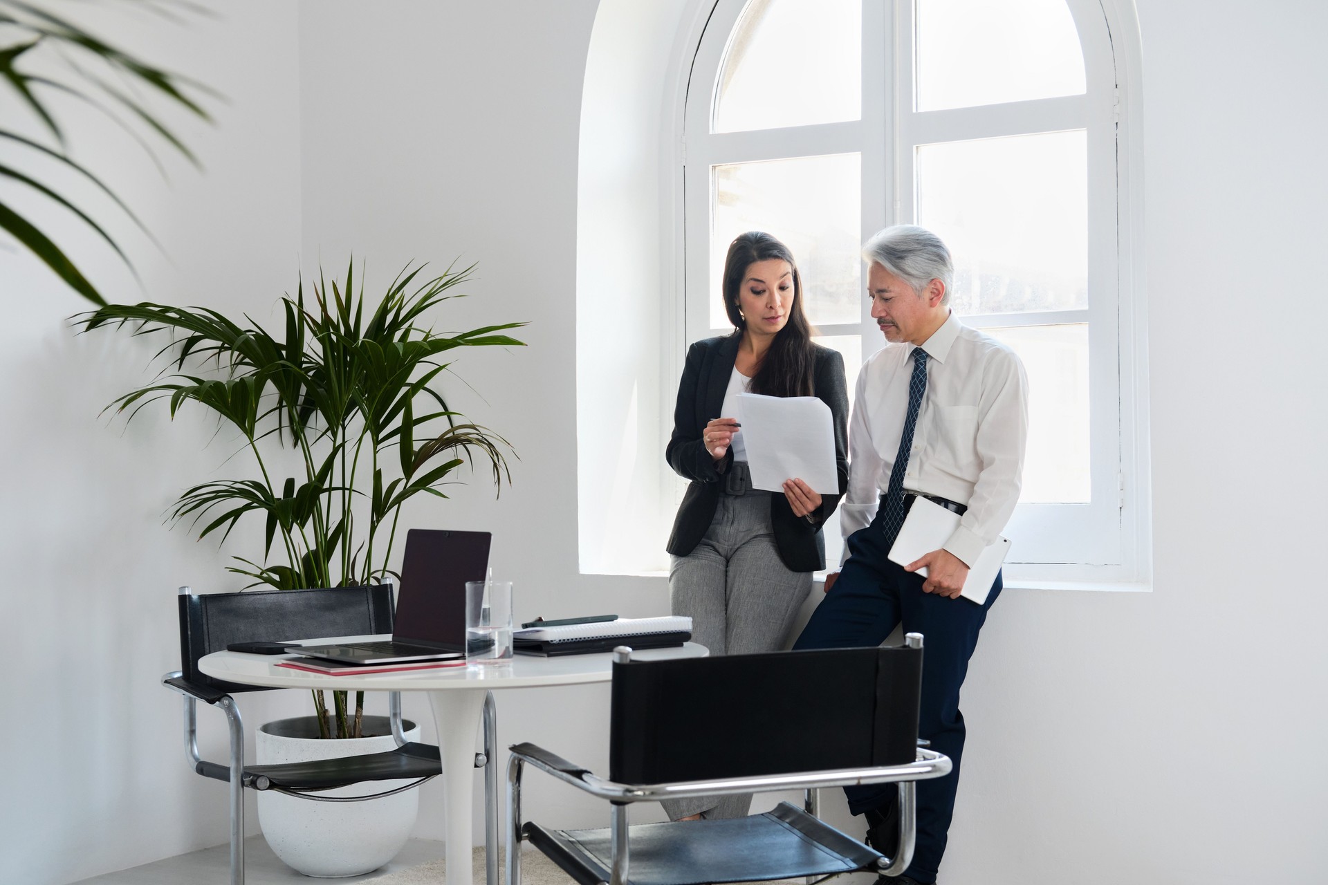 general shot and side view of a Japanese senior businessman and beautiful CEO businesswoman standing near a window focused looking at documents and talking before a business meeting in a cozy and lightly meeting room.